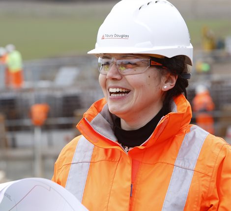 Female wearing an orange hi-vis jacket and a white hard hat, smiling while looking at paper plans, with the A602 road improvement works in the background