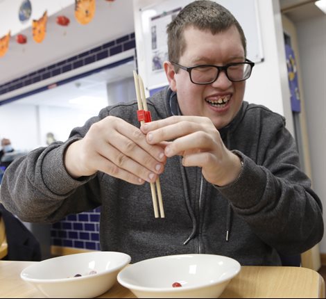 Male with glasses, wearing a grey hooded jumper, sat at a table smiling while using chopsticks to pick up and move multi-coloured sweets between two white bowls