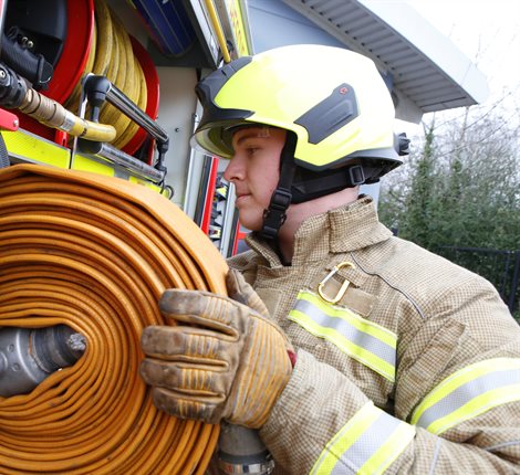 Male firefighter, wearing full personal, protective equipment, as he holds on to a yellow coiled hose which he is unloading from the side of a red fire engine