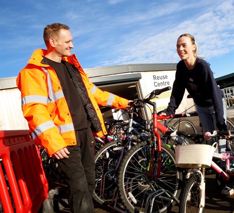 Male in orange hi-vis jacket stood showing bikes to a young female resident in a navy jumper and blue jeans, outside the front of Ware Recycle Centre on a sunny day with blue skies
