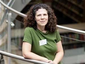 Person with curly hair wearing a green top and a name badge reading &ldquo;Herts &ndash; Abigail Lagden, Artist,&rdquo; leaning on a glass-and-metal railing inside Welwyn Garden City Library.