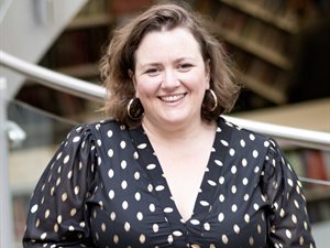 Close-up portrait of a person with short, wavy hair wearing a black blouse with gold polka dots and large hoop earrings, standing and resting crossed hands on a metal railing inside Welwyn Garden City Library.