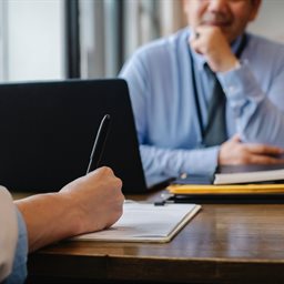 Woman writing at desk with man looking on