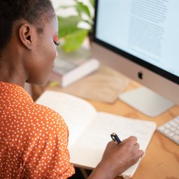 Woman in orange dress writing in a notebook in front of a computer